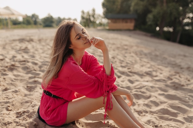 Aantrekkelijke stijlvolle Europese vrouw in roze jurk alleen zittend op het strand en genieten van recreatie in zonlicht