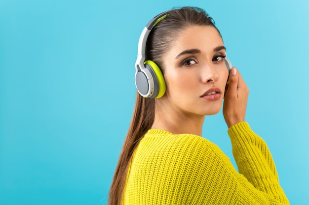 Aantrekkelijke stijlvolle jonge vrouw die naar muziek luistert in draadloze koptelefoons die blij zijn met het dragen van gele gebreide trui in kleurrijke stijl mode poseren