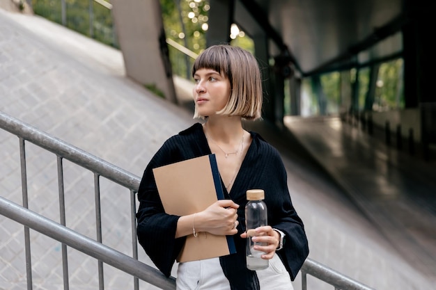 Gratis foto aantrekkelijke stijlvolle kortharige vrouw in blouse staat in het zakendistrict gelukkig meisje met steil kapsel met notitieboekjes en water buiten