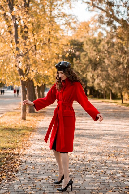 Aantrekkelijke stijlvolle lachende magere vrouw met krullend haar wandelen in het park gekleed in warme rode jas herfst trendy mode, Streetstyle, baret hoed dragen