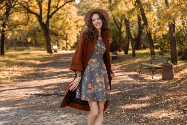 Aantrekkelijke stijlvolle lachende vrouw met krullend haar wandelen in park gekleed in gedrukte jurk en warme vacht herfst trendy mode, Streetstyle