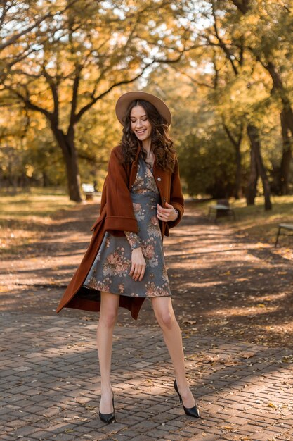 Aantrekkelijke stijlvolle lachende vrouw met krullend haar wandelen in park gekleed in gedrukte jurk en warme vacht herfst trendy mode, Streetstyle