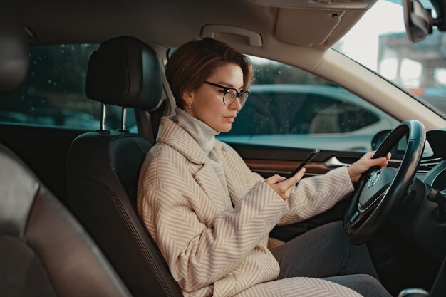 Aantrekkelijke stijlvolle vrouw zittend in de auto gekleed in jas winter stijl en bril met behulp van slimme telefoon