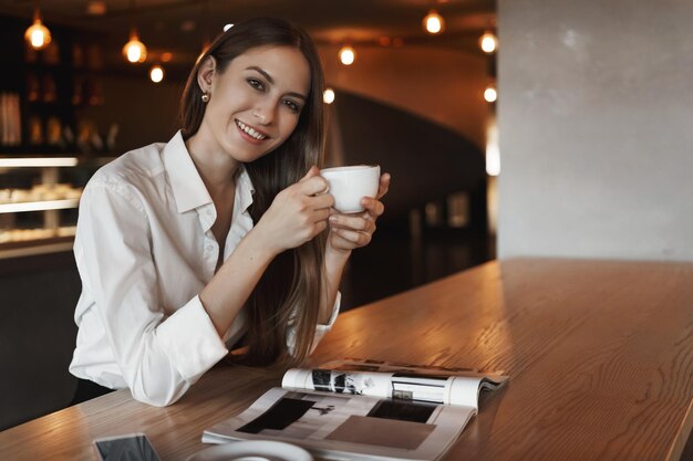 Aantrekkelijke succesvolle jonge vrouwelijke ondernemer die koffie drinkt in café en geniet van een leuk gesprek met een partner die een beker vasthoudt en een glimlachende camera kantelt hoofd van het lezen van modetijdschrift