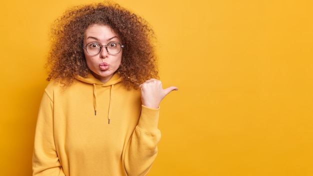 Aantrekkelijke verraste vrouw met krullend haar houdt lippen afgeronde punten met duim weg, maakt haar keuze bij het winkelen gekleed in casual hoodie poses over gele muur toont logo of promo-deal op kopieerruimte