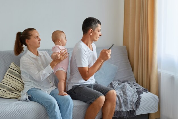 Aantrekkelijke vrouw die dochtertje in handen houdt, haar man achterover naar zijn familie zit en smartphone gebruikt, wil geen tijd doorbrengen met naaste mensen, surfen op internet of berichten typen.