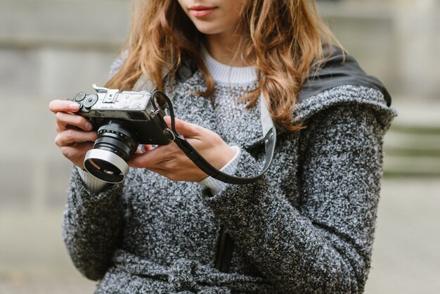 Aantrekkelijke vrouw die een grijze jas draagt die een uitstekende camera houdt en ernaar kijkt