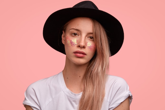 Aantrekkelijke vrouw hipster met heldere sparkles op wangen, draagt modieuze zwarte hoed, casual wit t-shirt, staat tegen roze muur