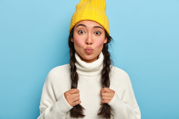 Aantrekkelijke vrouw houdt lippen rond, houdt twee staartjes, gekleed in witte warme trui en gele hoed, heeft flirterige blik op camera, geïsoleerd over blauwe muren