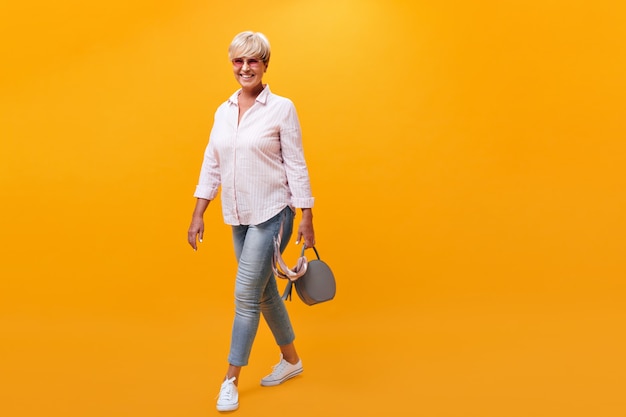 Aantrekkelijke vrouw in denim outfit beweegt op oranje achtergrond en houdt handtas