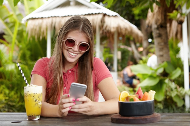 Aantrekkelijke vrouw in stijlvolle zonnebril slimme telefoon te houden en glimlachend met gelukkige blik terwijl ze bericht van haar vriend leest terwijl op vakantie in tropisch land.