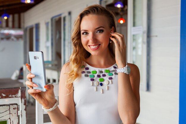 Aantrekkelijke vrouw in witte jurk in zomerterras in zonnebril met behulp van telefoon foto nemen