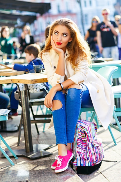 Aantrekkelijke vrouw met blond haar, sexy lachend meisje op terras, stad achtergrond. Een spijkerbroek met gaten, een witte jas.