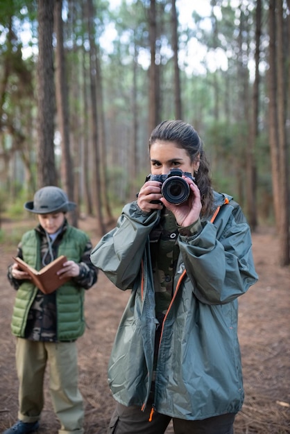 Aantrekkelijke vrouw met camera in bos. Vrouwelijk model in sportieve kleding glimlachend in de camera. Wazige jongen op de achtergrond. Hobby, fotografieconcept