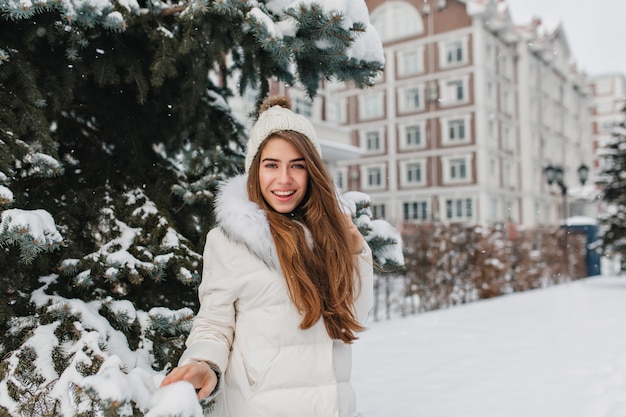 Aantrekkelijke vrouw met sluik bruin haar poseren met zelfverzekerde glimlach in de buurt van groene sparren in de winter. Prachtige jonge dame draagt een witte jas en een grappige hoed met plezier met sneeuw.