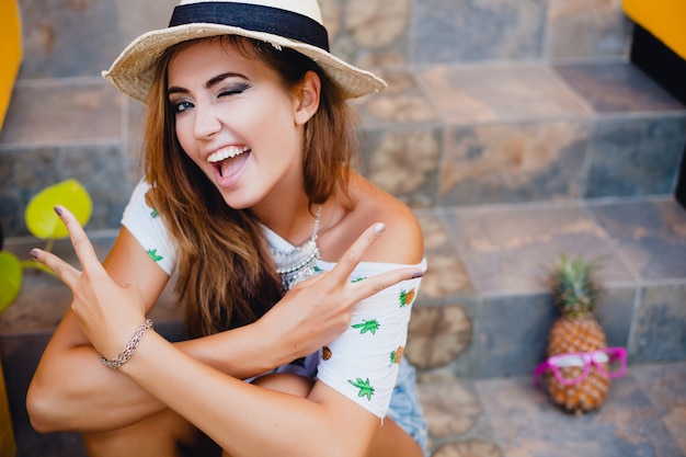 Aantrekkelijke vrouw op zomervakantie met grappige koele gezichtsuitdrukking die emotioneel glimlacht draagt strooien hoed knipogen