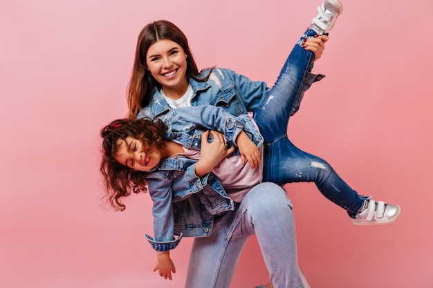 Aantrekkelijke vrouw speelt met dochtertje op roze achtergrond. studio shot van moeder en preteen kind in spijkerjasjes.