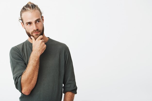 Aantrekkelijke Zweedse man met stijlvolle haren en baard in grijs shirt met zijn kin en nadenkend opzij kijken denken