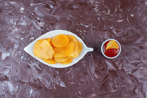 Gratis foto aardappelchips in een bord naast ketchup in een kom, op de marmeren tafel.