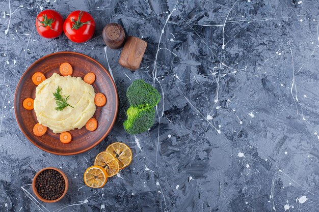 Aardappelpuree en gesneden wortelen op een bord naast groenten en kruidenkommen, op de blauwe tafel.