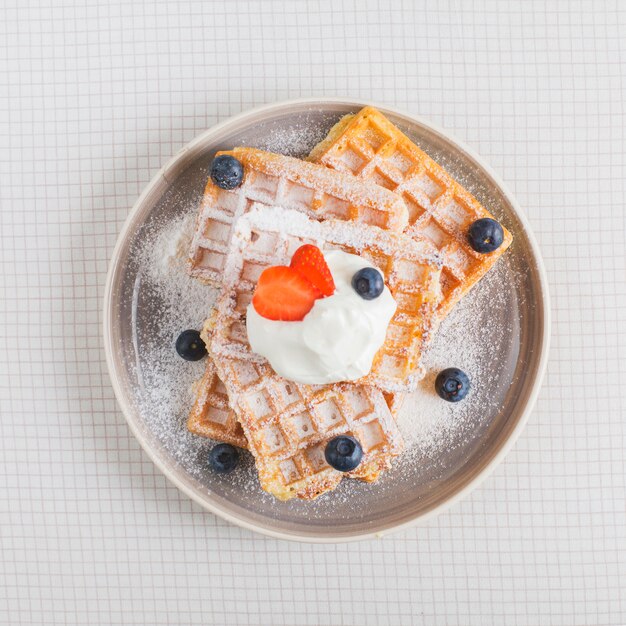 Aardbei en bosbes op slagroom over de stapel wafels op plaat over de geruite achtergrond