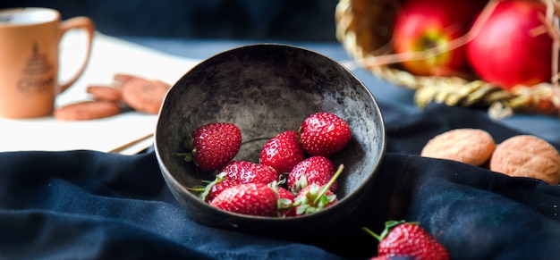 Aardbeien in de kom, koekjes, beker en appelmand op een zwarte mat.