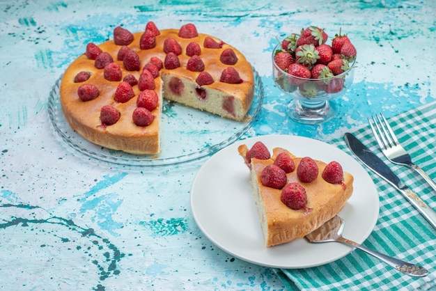 Gratis foto aardbeientaart gesneden en hele heerlijke cake samen met verse rode aardbeien op helderblauw bureau