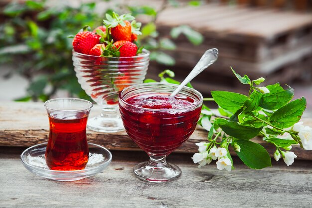 Aardbeijam in een plaat met lepel, thee in glas, aardbeien, installatie zijaanzicht op houten en werflijst