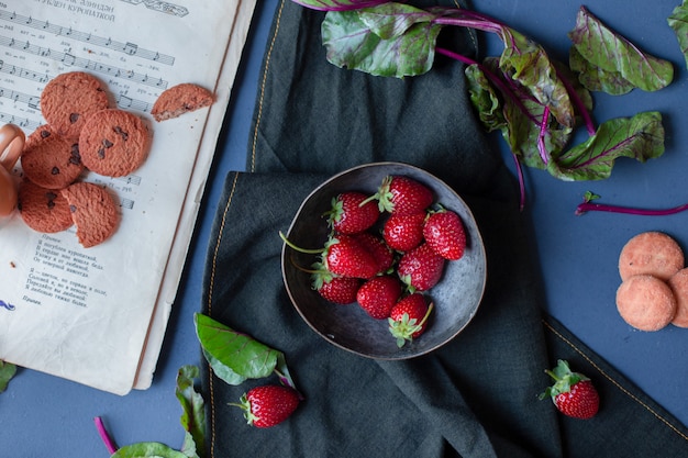 Aardbeikom en koekjes, spinaziebladeren, een boek rond op een zwarte mat.