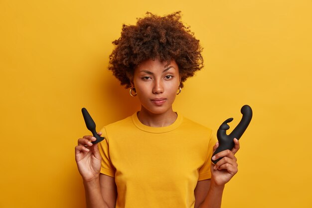 Aarzelende Afro-Amerikaanse vrouw houdt buttplug vast om op te warmen voor penetrerend spel, konijnvormige vibrator voor vagina stimulatie, gekleed in geel t-shirt. Jonge vrouw met seksspeeltjes binnen