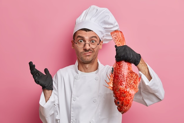 Aarzelende mannelijke chef-kok gekleed in uniform en hoed, zwarte handschoenen, houdt rode zeebaars, weet niet wat te koken, werkt in restaurant