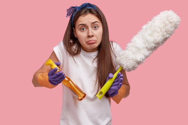 Aarzelende, ontevreden vrouw houdt stofdoek en schoonmaakvloeistof of spray vast, voelt apathie, wil de kamer niet opruimen