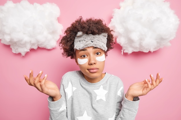 Aarzelende vrouw met krullend haar verspreidt handpalmen voelt twijfel gekleed in nachtkleding past collageenvlekken toe onder de ogen om donkere kringen te verwijderen draagt nachtkleding geïsoleerd over roze muur met wolken erboven