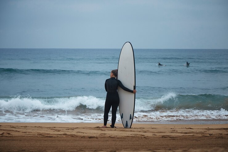 Gratis Foto achteraanzicht op mooie jonge surf meisje haar longboard knuffelen op de kust van de oceaan en kijken naar golven voor het surfen