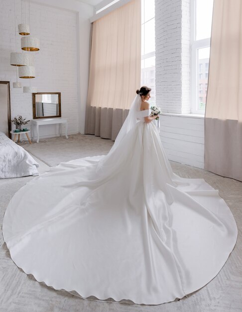 Achteraanzicht van aantrekkelijke bruid gekleed in luxe trouwjurk die in de kamer naast het raam staat