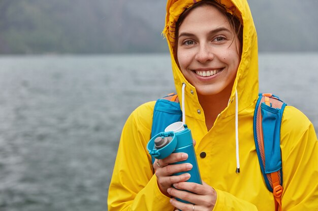 Achteraanzicht van blije vrouw draagt gele regenjas met capuchon, glimlacht gelukkig, wandelingen aan de oever van het bergmeer