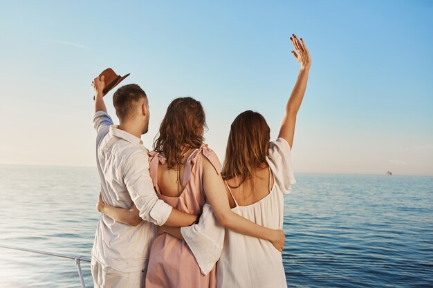 Achteraanzicht van drie beste vrienden reizen per boot knuffelen en zwaaien terwijl u op zee kijkt. Mensen die op luxe vakantie zijn, zeggen hallo tegen crue die per jacht passeert.