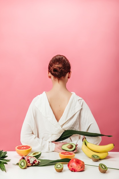 Gratis foto achteraanzicht van gember vrouw met exotisch fruit. studio shot van meisje in wit overhemd met bananen, grapefruits, granaat. kiwi en avocado op roze achtergrond.