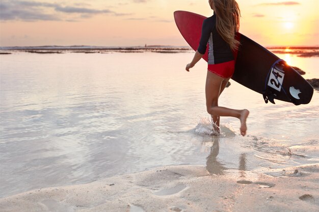 Achteraanzicht van het runnen van meisje surfer in zwembroek, draagt bord onder de arm, klaar om gigantische golf te veroveren, loopt in de oceaan