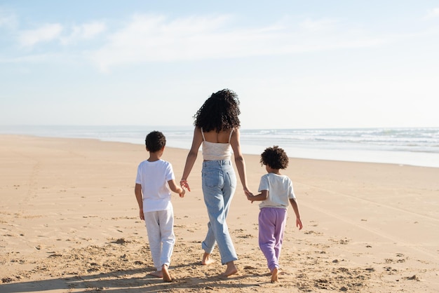 Achteraanzicht van moeder en kinderen hand in hand op het strand. afro-amerikaanse familie tijd samen doorbrengen in de open lucht. vrije tijd, familietijd, saamhorigheidsconcept