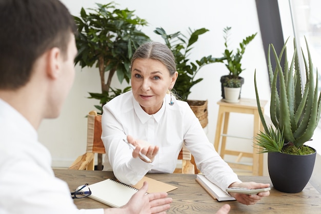 Achteraanzicht van onherkenbare jonge mannelijke kandidaat die een sollicitatiegesprek heeft met zelfverzekerde grijze haren recruiter van middelbare leeftijd die potlood en rekenmachine vasthoudt en hem vertelt over de gewenste positie