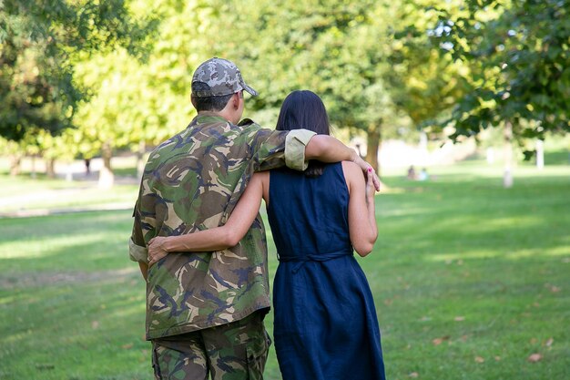 Achteraanzicht van paar knuffelen en samen wandelen op gazon in park. Man met camouflage uniform, zijn vrouw omhelzen en genieten van zonnige dag. Familiereünie, weekend en thuiskomst concept