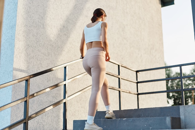 Achteraanzicht van slanke vrouw met een goede vorm die stijlvolle sportieve kleding, beige legging en top draagt, naar boven gaat om buiten te trainen, gezonde levensstijl, damestraining.