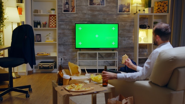 Achteraanzicht van zakenman die van zijn pizza geniet terwijl hij naar een tv kijkt met Mock Up groen scherm