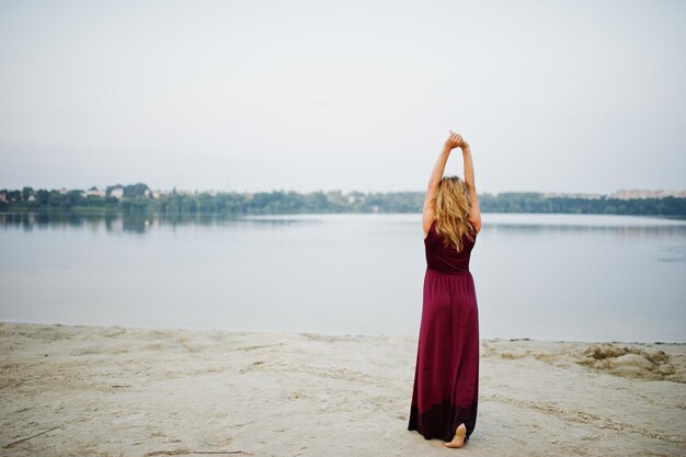 Achterkant van blonde sensuele barefoot vrouw in rode marsala jurk poseren tegen lake