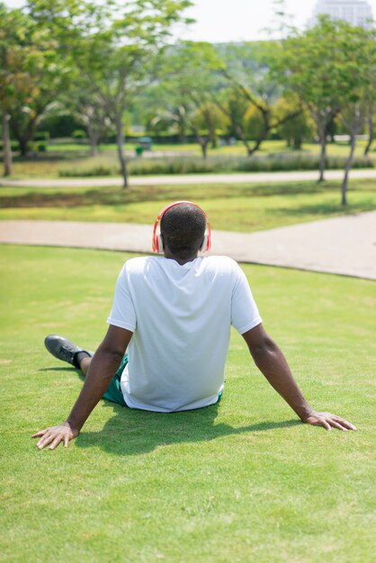 Achtermening van de zwarte mens die op gras met draadloze hoofdtelefoons rusten.
