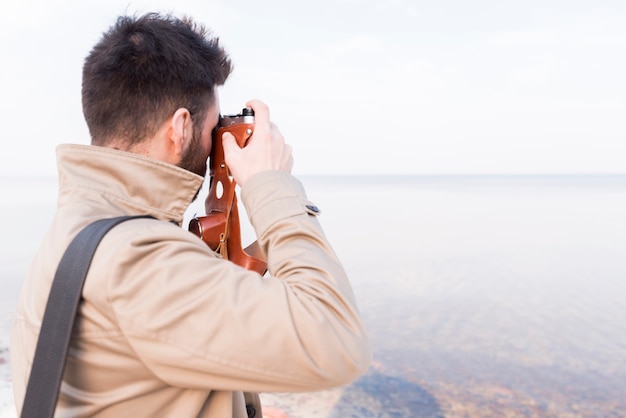 Gratis foto achtermening van een mannelijke reiziger die beeld van idyllische overzees met camera nemen