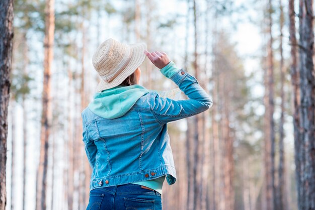 Achtermening van een vrouw die hoed op hoofd dragen die bomen in het bos bekijken