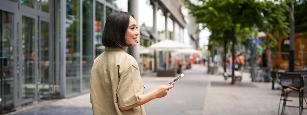 Gratis foto achterste opname van een jonge vrouw die door de stad loopt en door de straat loopt en glimlacht met smartphone-weergave