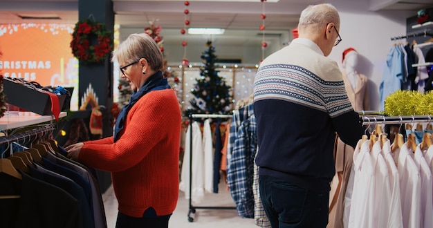 Gratis foto achtjarige klanten snuffelen tijdens de wintervakantie door kledingrekken in een feestelijke sierlijke modewinkel. senior koppel blij na het vinden van kleurrijke blazers om cadeau te doen tijdens kerstfamiliebijeenkomst